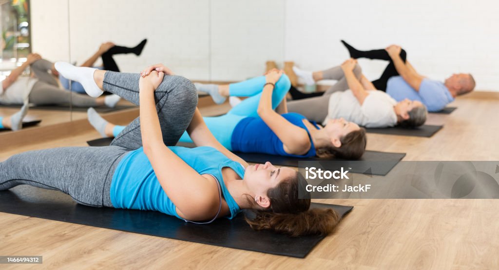 People of different ages stretching their glutes while lying on mats during Pilates workout in gym Acitve male and female of different ages lying on mats during Pilates stretching training in fitness studio 20-24 Years Stock Photo