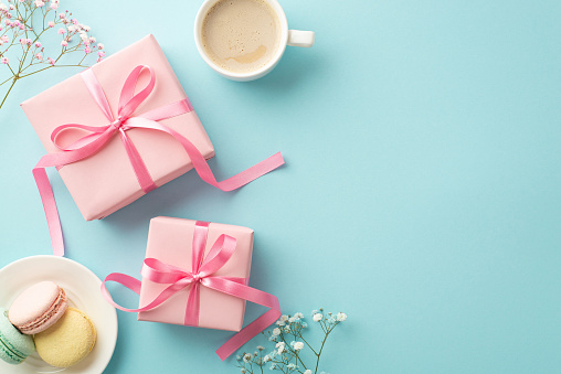 Mother's Day concept. Top view photo of pink gift boxes with bows saucer with macarons mug of fresh coffee and gypsophila flowers on isolated light blue background with copyspace