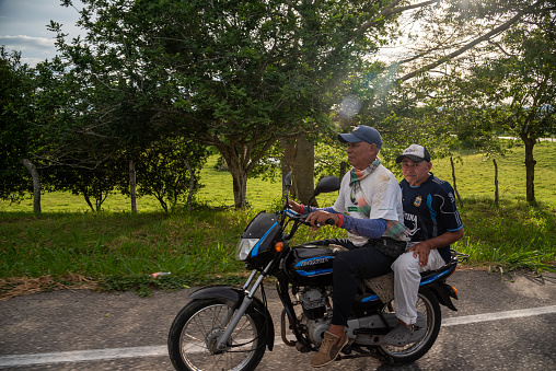 Two senior men on a motorbike on a country road at sunset time. Bolivar Department. Colombia. June 2, 2022.