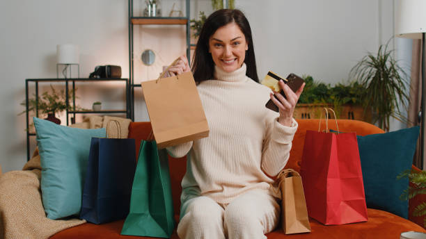 happy shopaholic jeune fille assise avec des sacs à provisions faisant le paiement en ligne avec une carte de crédit - women holding shopping bag living room photos et images de collection