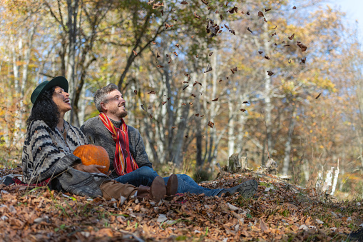 A beautiful Latin American woman and a Caucasian man, happy, in love and smiling, are sitting on the edge of the forest and having fun on an autumn trip. The man has a red scarf. They are enjoying the falling leaves, with a large Halloween pumpkin in their arms.