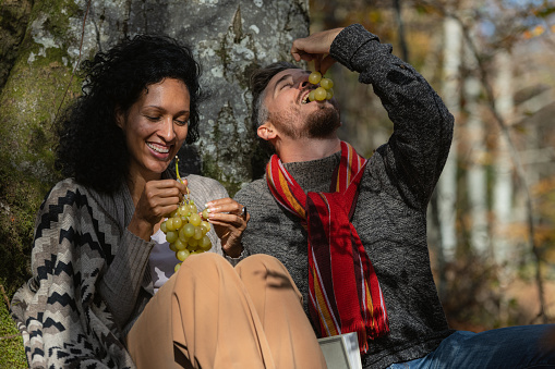 A beautiful Latina woman and a white man, happy, in love and smiling, have fun on an autumn trip in the forest and eat grapes.
The man has a red scarf.