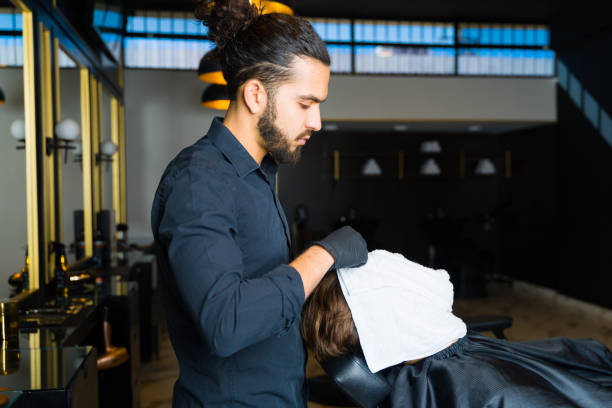 Professional hispanic barber putting a hot towel on a relaxed young man to help the skin before shaving his beard