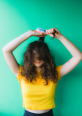 Young woman in yellow t-shirt trying to tame her fizzy curly hair with brush on green background