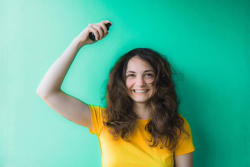 Young woman in yellow t-shirt trying to tame her frizzy curly hair and spraying hair care on it  on green background
