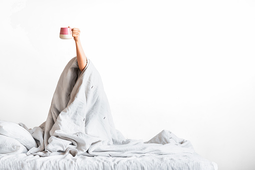 Raised female hand with cup of coffee - young woman with cup of coffee sitting in bed under blanket at home bedroom.