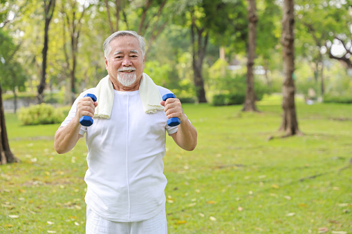 Happy and smiling asian senior man doing arm work out and lifting dumbbell exercise with relaxation for healthy in park outdoor after retirement. Health care elderly outdoor lifestyle concept.