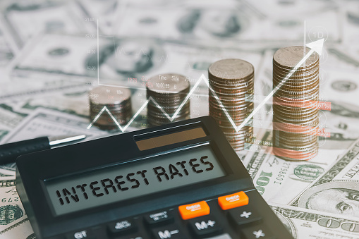 The word interest rate is on a calculator with a coin, a graph, and an arrow pointing up.  Concepts of raising interest rates, economic conditions, inflation, etc.