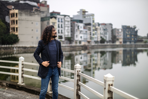 Young man with long curly hair and beard, wearing an suit jacket,  standing next to the urban lake with in Long Bien, Hanoi, Vietnam