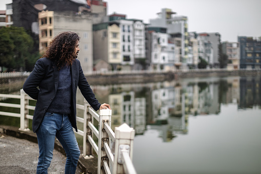 Young man with long curly hair and beard, wearing an suit jacket,  standing next to the urban lake with in Long Bien, Hanoi, Vietnam