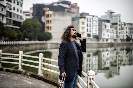 Young man with long curly hair and beard, wearing an suit jacket,  standing next to the urban lake with pink suitcase and backpack in Long Bien, Hanoi, Vietnam