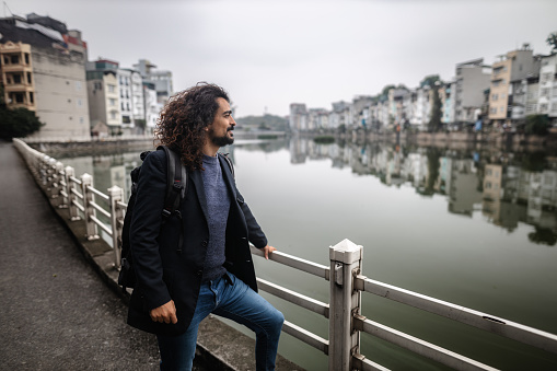 Young man with long curly hair and beard, wearing an suit jacket,  looking at the urban lake, carrying a backpack in Long Bien, Hanoi, Vietnam