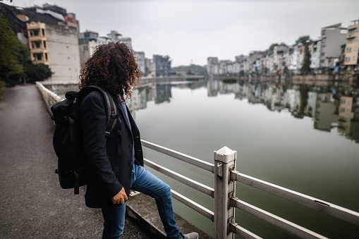 Young man with long curly hair and beard, wearing an suit jacket,  looking at the urban lake, carrying a backpack in Long Bien, Hanoi, Vietnam