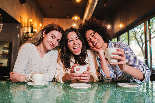 Three young women smiling looking at camera in a coffee shop. A group of multiracial happy girls having fun together taking a break in a restaurant. High quality photo