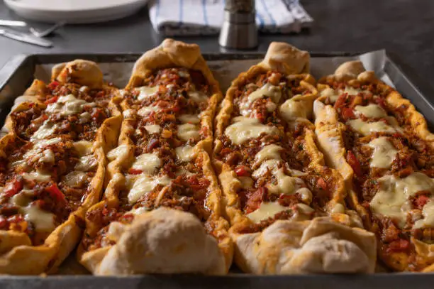 Delicious homemade turkish pide or savory pastry boats with minced meat, tomatoes, peppers, onions, garlic and herbs. Topped with melted cheese and served hot on a baking sheet on kitchen table. Closeup and front view