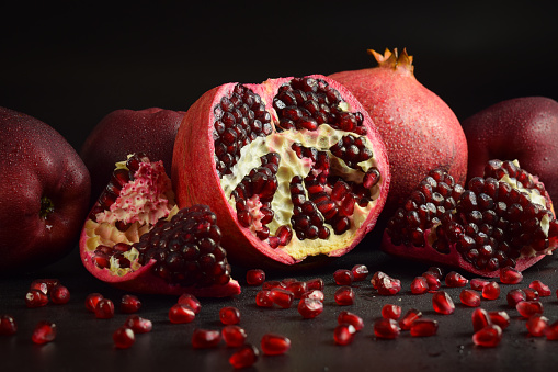 Pomegranate clusters with fresh pomegranate arils. Still life ripe pomegranates, red apples, green leaves on black background. Split open fruit scattered red grains seed indoors. Studio shot