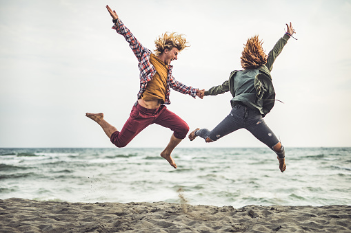 Young carefree couple having fun while holding hands and jumping on the beach in autumn day. Copy space.