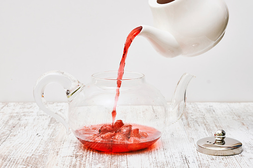A teapot pouring strawberry tea into a glass pot on a wooden table against a white wall