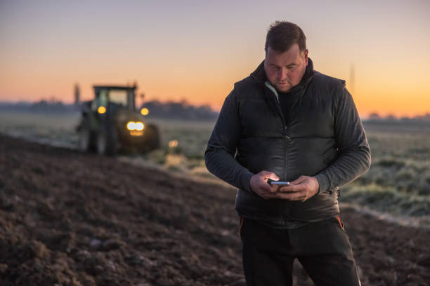 istock Man with short brown hair looking down while using his smartphone,standing on an agricultural field in the evening,tractor with lights in the background 1466631454