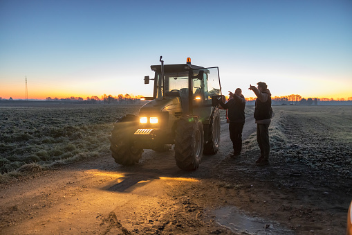 Two male farmer standing next to a green tractor in the evening on an agricultural path,pointing in a direction with their finger while talking,lights on the vehicle are on