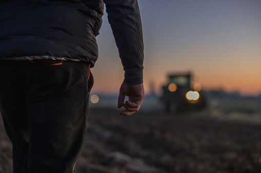 Close-up male farmer wearing trousers and a black vest standing on his agricultural field,green tractor with lights turned on in the background,focus on foreground
