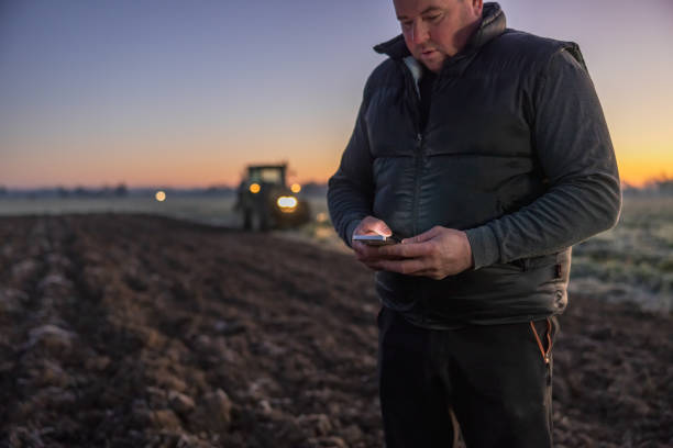 istock Male farmer looking down on his smartphone with worried facial expression while standing on his agricultural field,tractor with lights on in the background 1466632239