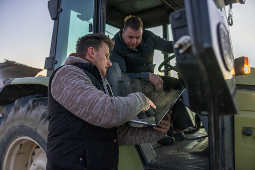 Man with short brown hair,wearing a gray pullover and black vest pointing on his laptop while standing next to his colleague,colleague sitting inside his green tractor and looking on the screen,in the evening