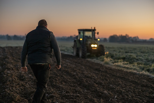 Hombre con cabello castaño corto, y un chaleco negro está caminando en su campo agrícola por la noche, con tractor en el fondo, las luces del tractor están encendidas photo