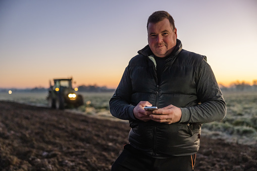 Male farmer with short brown hair smiling and looking at camera while using his smartphone,standing on an agricultural field in the evening,green tractor with lights turned on in the background