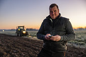 Male farmer with short brown hair smiling and looking at camera while using his smartphone,standing on an agricultural field in the evening,tractor with lights in the background