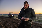 Male farmer with short brown hair writing a message on his smartphone while standing on his agricultural field in the evening,tractor with lights in the background