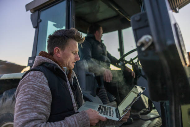 Man with short brown hair using his laptop while standing next to a tractor with a male farmer sitting in it in the evening