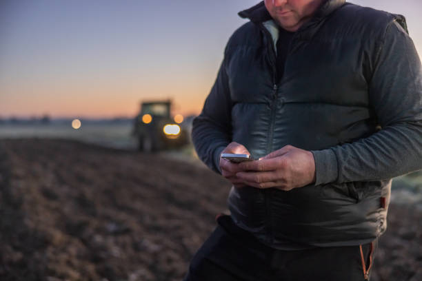 istock Man looking down on his smartphone with while standing on his agricultural field,tractor with lights on in the background,sunset at the horizon 1466632968