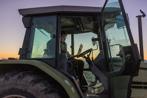 Man with short brown hair,wearing a gray pullover,trousers and black vest is sitting inside of his green tractor,working on his laptop,side view,in the evening