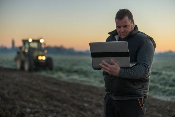 istock Man with short brown hair is using his laptop while standing on an agricultural field during sunset,tractor with lights in the background 1466635445