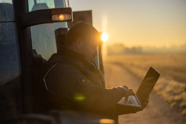 istock Man with short brown hair is using his laptop while leaning against his tractor in the evening,sunbeam from the back,in silhouette 1466635452
