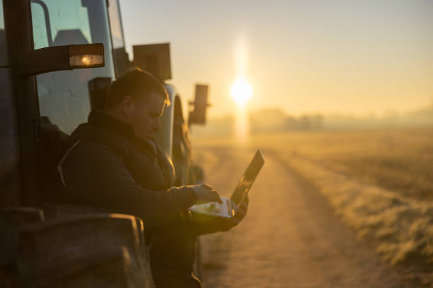 istock Male farmer with short brown hair is analyzing the agricultural processes on his laptop while leaning against his tractor in the evening 1466635663
