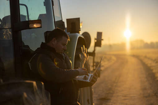 Male farmer with short brown hair,wearing a gray pullover and black vest is working on his laptop while leaning against his green tractor in the evening,sunset in the background,side view