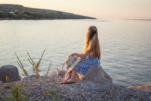 Girl with long blond hair sits on a stone on the banks of a river, lake, sea. She is looking at ocean and thinking dreamily. Girl alone outside. Girl sitting on rocks. Lonely person. Vacation on sea. Girl thinking on the beach.