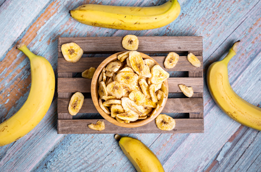 A top view of dehydrated organic banana chips on the wooden board, decorated with fresh bananas