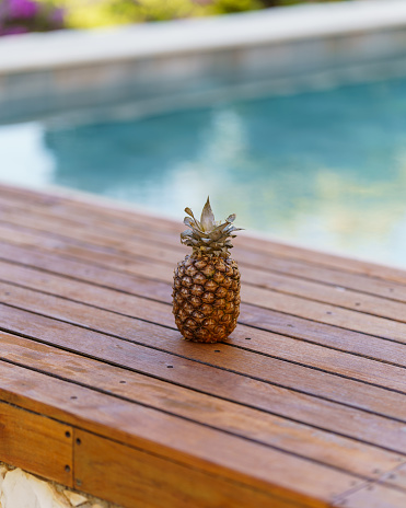 A vertical shot of a pineapple on a wooden bench near a pool
