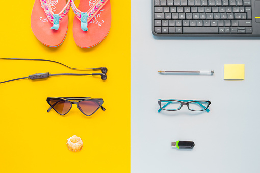 Flip-flops, sunglasses, seashell, keyboard, pen and headphones on a yellow and gray cardboard. Vacation and work concept. Top view, symmetry.