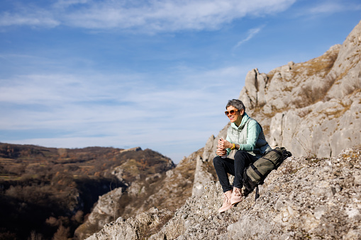 Mature woman sitting on rock and opening thermos while taking a break from hiking on rocky mountain