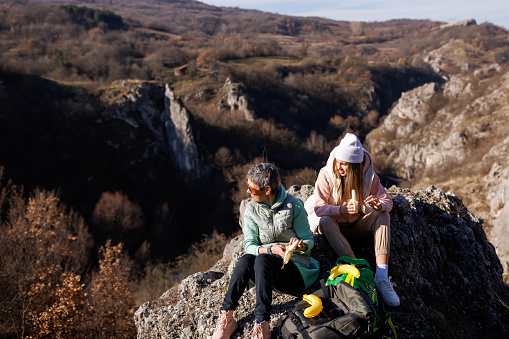 Mature mother and young adult daughter sitting on rock and eating banana after hiking on rocky mountain on sunny day