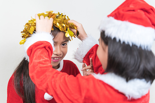 Little girl with Santa costume helping another girl to wear a head garland on a white background.
