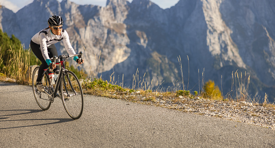 Female cyclist with sports clothing and helmet riding her bike on a road next to the abyss in the beautiful mountain scenery at Mangart during sunrise