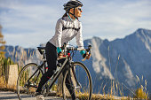 Adult woman with brown hair taking a break from cycling,standing on a road with her bike and looking at the view of the Mangart mountain scenery