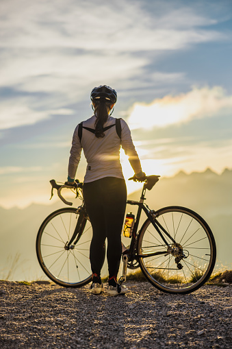 Adult woman with long brown hair tied in a ponytail,wearing sports clothing,standing on mountain peak during sunrise with her bike,looking at the beautiful view,rear view