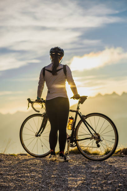 Adult woman with long brown hair standing on mountain peak during sunrise with her bike,looking at the view Adult woman with long brown hair tied in a ponytail,wearing sports clothing,standing on mountain peak during sunrise with her bike,looking at the beautiful view,rear view female mountain biker silhouette stock pictures, royalty-free photos & images
