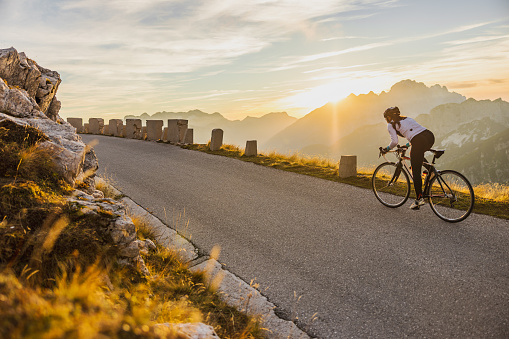 Woman with long brown hair tied in a ponytail,wearing helmet and sports clothing riding her bike up a tight serpentine road in the Mangart mountain scenery during sunrise